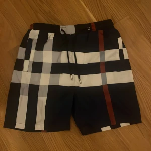 Rutiga  Bad Burberry shorts med snörnin - Snygga Badshorts från Burberry med klassiskt rutigt mönster i svart, vitt, grått och röda detaljer. De har elastisk midja med snörning och sidofickor. Perfekta för sommaren när du vill sticka ut med en lyxig och trendig look.