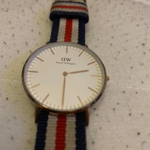 Daniel Wellington klocka med randigt band - Stilren klocka från Daniel Wellington med vit urtavla och guldfärgad boett. Bandet är randigt i marinblått, rött och vitt, tillverkat i textil. Minimalistisk design med tunna visare och indexmarkeringar. Perfekt accessoar för en clean och klassisk look. Fungerar bra