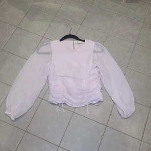 Ljuslila blus med puffärmar från Lindex - Supersöt ljuslila blus från Lindex med transparenta puffärmar och snygga snörningsdetaljer i sidorna. Blusen har rund halsringning, knapp i nacken och smock baktill för extra komfort. Perfekt för dig som gillar romantisk och trendig stil.