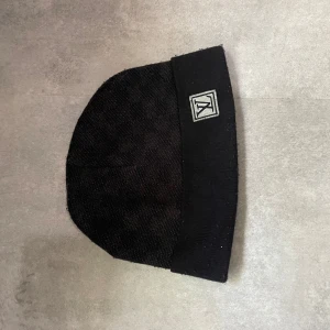 Svart mössa från Louis Vuitton - Snygg svart mössa från Louis Vuitton med diskret LV-logga framtill. Klassisk form med uppvikt kant och vävd struktur. Perfekt accessoar för dig som gillar exklusiva märken och stilren design. Använd 3-4 gånger.