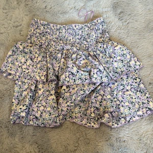 Blommig volangkjol från Gina Tricot - Supersöt kort volangkjol från Gina Tricot med blommigt mönster i lila, gul, svart och vit. Kjolen har smock i midjan och flera lager volanger 