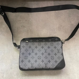 Louis Vuitton svart axelväska monogram - Snygg svart axelväska från Louis Vuitton med klassiskt monogrammönster i grått. Väskan har justerbar axelrem, silverfärgade dragkedjor och är tillverkad i skinn. Perfekt storlek för att bära det viktigaste och har flera fack inuti.
