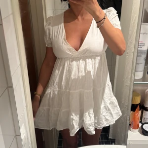 Vit kort klänning med v-ringning - Supersöt vit kort klänning med djup v-ringning och puffiga korta ärmar. Klänningen har volangkant nertill och är sydd i ett lätt, luftigt bomullstyg.