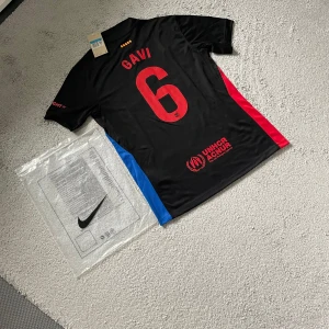 FC Barcelona Gavi fotbollströja Nike - Snygg svart FC Barcelona fotbollströja från Nike med Gavi och nummer 6 på ryggen. Tröjan har röda och blå sidopaneler, klubbmärke på bröstet och UNHCR-logga. Materialet är lätt och andas, perfekt för match eller träning. Kortärmad modell med modern passform.