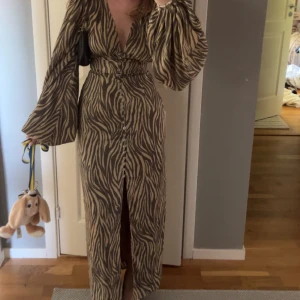Adoore klänning  - Snygg långklänning med zebramönster i beige och brunt från adoore. Djup v-ringning, vida långa ärmar och knappar framtill hela vägen ner. Klänningen har en slits framtill och markerad midja för en riktigt trendig siluett. Perfekt för dig som vill sticka ut! Den har en liten defekt som man ser på sista bilden men inget som läggs märke till och går att fixa då snöret har gått av