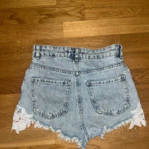 Högmidjade jeansshorts med spets - Snygga ljusblå jeansshorts med hög midja och råa, fransiga kanter. Shortsen har vita spetsdetaljer längs bensluten som ger en unik och trendig look. Klassiska fickor fram och bak samt bälteshällor. Perfekta för sommarens alla äventyr!