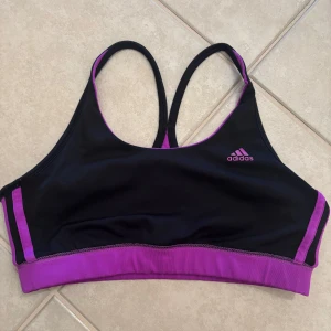 Svart och lila sporttopp från Adidas - Sportig topp från Adidas i svart och lila med klassiska loggan framtill. Snygga kontrastdetaljer och smala axelband som korsas i ryggen. Tillverkad i stretchigt syntetmaterial som andas, perfekt för träning och aktiva dagar.