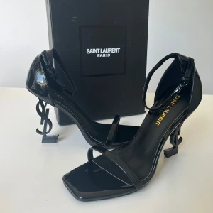 Svarta Saint Laurent klackar med YSL-häl - Superstilrena svarta klackar från Saint Laurent i lackat skinn med ikonisk YSL-logga som klack. Skorna har fyrkantig tå, smala remmar över foten och runt ankeln med spänne. Perfekta för dig som vill sticka ut med en lyxig och edgy vibe.