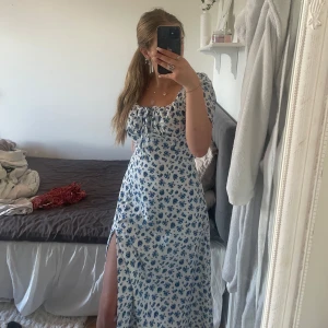 Blommig långklänning med slits - Superfin långklänning i vitt med blått blommönster, puffärmar och söt snörning vid bysten. Klänningen har en snygg slits på sidan!