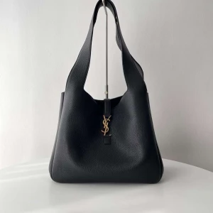 Svart axelväska från Saint Laurent - Säljer en svart axelväska från Saint Laurent i exklusivt skinn med guldigt YSL-emblem framtill. Väskan har en stilren, rymlig design med bred axelrem och innerfack med dragkedja. Perfekt för dig som vill ha en lyxig och tidlös accessoar.