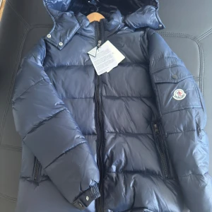 Mörkblå Moncler dunjacka med huva - Snygg mörkblå dunjacka från Moncler med huva och dragkedja framtill. Jackan har Moncler-logga på ärmen, praktiska fickor med dragkedja och quiltad design. Perfekt inför hösten till ett grymt pris så passa på! QR-scannern fungerar som den ska och jackan är i ny skick med lappen kvar på😊