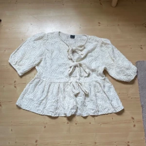 Beige blus med knyt och struktur - Supersöt beige blus från Gina Tricot med tre knyt framtill och ett vågigt strukturerat tyg. Blusen har v-ringning, volang nertill och halvlånga ärmar. 