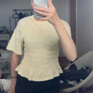 Gul volangtopp från Gina Tricot - Supersöt ljusgul topp från Gina Tricot med smockad midja och volang nertill. Toppen har korta ärmar och en figurnära passform över kroppen. Perfekt för dig som gillar en feminin och trendig stil.