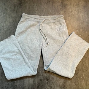 Grå mjukisbyxor från GAP med snörning - Snygga ljusgrå mjukisbyxor från GAP med broderad logga på ena benet. Byxorna har dragsko i midjan för perfekt passform och är gjorda i mjukt bomullsmaterial. Perfekta för chill dagar eller när du vill vara extra bekväm.