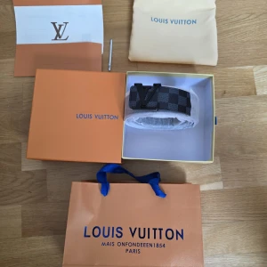 Louis Vuitton rutigt bälte svart - Snyggt bälte från Louis Vuitton med klassiskt rutigt mönster i svart och grått. Bältet har en stor LV-logga i svart metall som spänne och är gjort i exklusivt skinn. Perfekt accessoar för att lyfta din outfit.