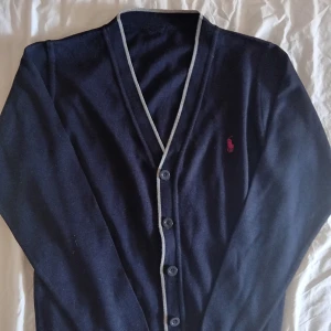 Mörkblå kofta från Polo Ralph Lauren - Snygg mörkblå kofta från Polo Ralph Lauren med klassisk röd broderad logga på bröstet. Koftan har v-ringning, knäppning framtill med märkta knappar och ljus kontrastkant längs öppningen. Tillverkad i mjuk bomull, perfekt för lager-på-lager under höst och vår.(tagen saknas)