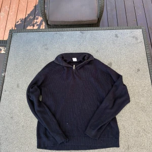 Mörkblå stickad tröja med half zip - Mörkblå stickad tröja med half zip och hög krage. Tröjan har lång ärm och ribbade muddar vid ärmslut och nederkant. Perfekt för kyliga dagar och enkel att matcha med jeans eller chinos.