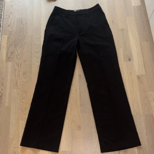 Svarta kostymbyxor från Madlady - Svarta kostymbyxor från Madlady i klassisk rak modell med normal passform. Byxorna har hög midja, pressveck och sidfickor. Perfekta för en clean och stilren look. Materialet är mjukt och följsamt, vilket gör dem bekväma hela dagen.