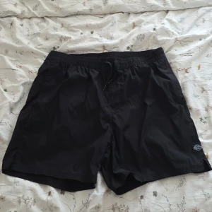 Svarta badshorts från FSBN XL - Svarta badshorts från FSBN i storlek XL. De har elastisk midja med snörning och en bakficka med dragkedja. Perfekta för stranden eller poolen, tillverkade i lätt syntetmaterial som torkar snabbt. Enkel och stilren design utan mönster.