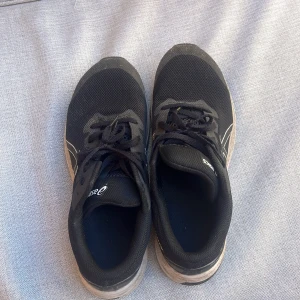 Svarta Asics sneakers strl 40 - Svarta Asics sneakers med vit logga och detaljer. Ovandel i mesh för bra andningsförmåga och snörning framtill. Sulan är svart och vit med bra grepp, perfekt för träning eller långa promenader. Klassisk sportig look och bekväm passform.