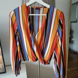 Randig omlottblus från Gina Tricot XS - Färgstark omlottblus från Gina Tricot i storlek XS. Blusen har långa ärmar med resår vid ärmslut och ett snyggt omlottliv. Mönstret är randigt i orange, blått, svart, vitt och gult. 