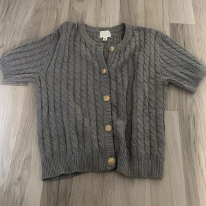 Grå kabelstickad kofta med kort ärm - Grå kabelstickad kofta från H&M med korta ärmar och rund hals. Koftan har snygga träfärgade knappar framtill och ribbade muddar vid ärmslut och nederkant. Perfekt att slänga över en t-shirt för en chill look. Precis som med den vinröda så köpte jag i M för att S va lite för kort, pris kan alltid diskuteras