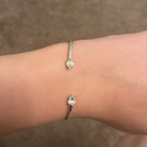 Caroline Svedbom armand silver med genomskinliga stenar - Stilrent armband i silverfärg med två gnistrande stenar i varje ände. Armbandet är öppet och smalt, vilket ger en modern och elegant look. Perfekt för dig som gillar enkla men snygga accessoarer.