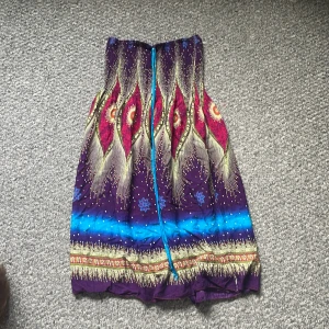 Färgglad kort klänning - Säljer en superfin kortklänning med färgstarkt påfågelfjädermönster i lila, blått, gult och rött. Klänningen har bandeau-modell utan axelband och en smockad överdel med knytband. Perfekt för sommarens varma dagar! 50kr plus frakt.