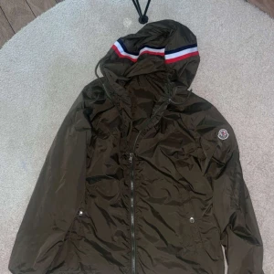Olivgrön vindjacka från Moncler - Snygg olivgrön vindjacka från Moncler med klassisk logga på ärmen. Jackan har dragsko i huvan och är tillverkad i glansig polyester som ger en clean och sportig look. Perfekt för dig som vill ha en lätt jacka med exklusiv känsla. Pris kan diskuteras vid snabbaffär!