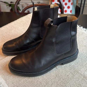 Svarta chelsea boots från Arket - Stilrena svarta chelsea boots från Arket i äkta skinn. Klassisk rund tå, elastiska paneler på sidorna och dragflikar fram och bak för enkel påtagning. Snygg, platt sula med robust känsla och diskreta sömmar. Perfekta för dig som gillar minimalistisk och tidlös stil.