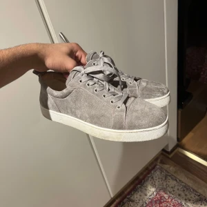 Grå mockasneakers från Christian Louboutin - Snygga sneakers i grå mocka från Christian Louboutin med klassisk röd sula och vita kanter. Skorna har rund tå, platt sula och snörning. Insidan är i beige skinn och designen är stilren men ändå lyxig. Perfekta för dig som gillar exklusiva detaljer. Skriv om ni har fler frågor! 