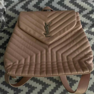 Beige quiltad ryggsäck från YSL - Säljer en snygg beige ryggsäck från YSL med ikoniskt metallogo framtill. Väskan är quiltad i ett mönster med tydliga sömmar och har justerbara axelremmar. Perfekt för dig som vill ha en lyxig och trendig accessoar till vardagen.