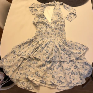 sommar klänning - Supersöt kortklänning med blå blommor på vit botten, omlott över bysten och knyt i nacken. Smockad midja och volangig kjol ger en romantisk vibe. Perfekt för varma dagar och sommarhäng. aldrig använd 