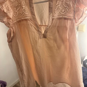 Beige blus med broderade detaljer - Säljer en luftig beige blus med v-ringning och broderade detaljer upptill. Blusen har korta, voluminösa ärmar och är tillverkad i ett tunt, lätt material som känns perfekt för varmare dagar. Broderierna ger en bohemisk touch och toppen är riktigt flowy.