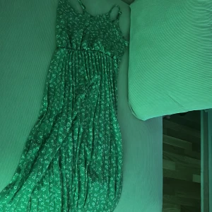 Grön blommig långklänning med smala axelband - Snygg långklänning i grönt med små vita blommor och smala axelband. Klänningen har en smockad midja och ett luftigt, plisserat fall som ger en avslappnad men ändå chic look. Perfekt för varma dagar när du vill känna dig både bekväm och trendig.