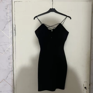 Svart kort klänning från NLY One - Snygg svart kort klänning från NLY One med tunna axelband och trendig snörning framtill. Klänningen har en figurnära passform och dragkedja bak. Perfekt för dig som gillar stilrena och enkla plagg med en twist.