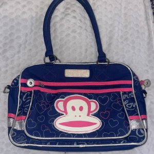 Paul Frank axelväska med hjärtan - Blå axelväska från Paul Frank med stort Julius-tryck framtill, rosa dragkedjor och silverdetaljer. Väskan har hjärtmönster, dubbla handtag och justerbar axelrem. Tillverkad i slitstark polyester med flera fack och innerficka med dragkedja. Finns tecken av användning💖