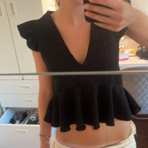 Svart blus med volang och v-ringning - Supersöt svart blus med djup v-ringning, volangdetaljer nertill och på axlarna. Ärmlös modell med broderade hål i tyget som ger en luftig känsla. 