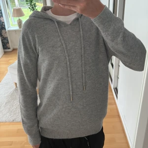 Grå stickad hoodie med huva och snörning - Mysig grå stickad hoodie med huva och långa ärmar. Tröjan har snörning i halsen och ribbade muddar vid ärmslut och nederkant. Perfekt för dig som gillar en enkel och stilren look med en chill vibe.