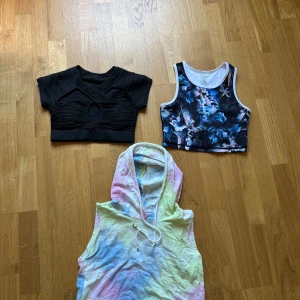 Tre trendiga croppade toppar - Säljer tre snygga croppade toppar: en svart ribbad topp med coola utskärningar, en sportig mönstrad topp i blått och vitt samt en ärmlös hoodie i pastellfärgad tie-dye. Perfekta för dig som gillar att sticka ut och mixa stilar.