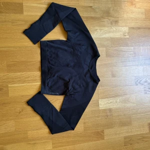 Svart croppad träningströja  - Svart croppad långärmad träningströja. Tröjan är i ett stretchigt syntetmaterial och har diskreta ribbade detaljer på ärmarna. Perfekt för gymmet eller löprundan och sitter tajt mot kroppen för en sportig look.