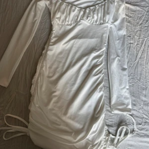 Vit långärmad klänning från Effordram - Supersnygg vit kortklänning från Effordram med långa ärmar och trendig snörning i sidorna. Klänningen har en lätt glansig finish och är figurnära med rynkade detaljer längs sidorna. Perfekt för dig som gillar stilrena och moderna plagg.
