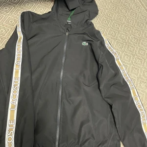 Svart tracksuit från Lacoste - Säljer en svart tracksuit från Lacoste med huva och dragkedja. Jackan har vita och beige ränder med Lacoste-logga längs ärmarna och klassisk krokodil på bröstet. Byxorna har grön piping och matchande krokodillogga. Perfekt för sportiga dagar. Helt ny inga skador äkta oanvänd finns i storlekarna M/L