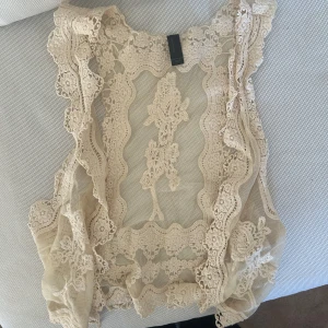 Beige spetsig bolerotopp  - Supersöt bolerotopp i beige spets. Toppens mesh är dekorerad med broderade blommor och vågiga kanter. Perfekt att slänga över ett linne eller klänning för en extra touch. Ärmlös och med romantiska volangdetaljer.