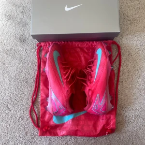 Nike Air Zoom Mercurial fotbollsskor - Säljer ett par Nike Air Zoom Mercurial fotbollsskor i en riktigt snygg röd färg med ljusblå detaljer och sula. Skorna har snörning och är tillverkade i syntetmaterial för bästa prestanda på planen. Perfekta för dig som vill sticka ut med stil och fart. Har använts två träningar och finns kvitto 