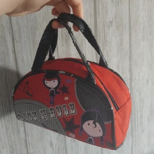 Röd handväska med rockmotiv - Cool röd handväska med svarta handtag och tryck av tecknade figurer, hjärtan och texten 'Rock N Rule' på framsidan. Väskan har dragkedja upptill och är tillverkad i tyg med detaljer i syntet. Perfekt för dig som gillar unika accessoarer med attityd.