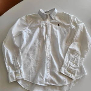 Vit skjorta från Ralph Lauren - Klassisk vit skjorta från Ralph Lauren i slim fit-modell. Skjortan är tillverkad i 100% bomull och har långa ärmar, knappar framtill och broderad logga på bröstet. Perfekt för en clean och stilren look.