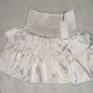 Vit satinvolangkjol från Neo Noir - Supersöt kort kjol i pärlvit satin från Neo Noir med prislapp kostar 600 kr helt ny. Kjolen har bred smock i midjan och dubbla volanger som ger en riktigt flowy och trendig look. Perfekt att styla med en croppad topp eller stickad tröja för en fräsch vibe.