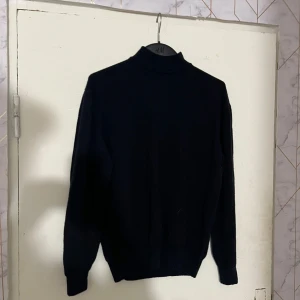 Svart polotröja i merinoull - Klassisk svart polotröja i ren merinoull från Woolmark. Tröjan har lång ärm och hög krage, perfekt för lager-på-lager under kyliga dagar. Mjuk och skön känsla mot huden, passar till både jeans och kostymbyxor.