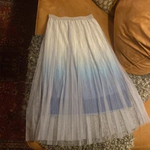 Glittrig långkjol från Ed London - Säljer en superfin långkjol från Ed London i glittrigt tyg. Kjolen är plisserad och har en snygg ombre-effekt i vitt och blått. Perfekt för dig som vill sticka ut med en drömmig och trendig look. Resår i midjan för extra komfort.